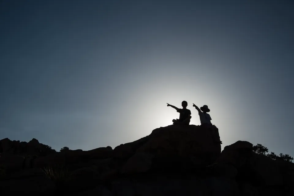 Two children sitting on rock sillhouetted against sky.
